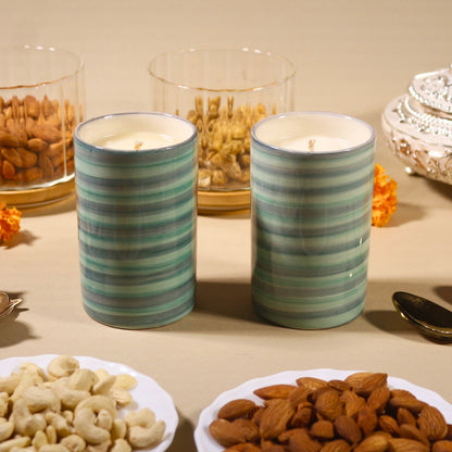 Two ceramic jar candles with green and white stripes, displayed on a table with various nuts and a decorative element in the background.