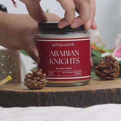 Whipandwick Arabian Knights soy wax candle in a red glass jar, lit and surrounded by deep red roses against a dark, elegant background.